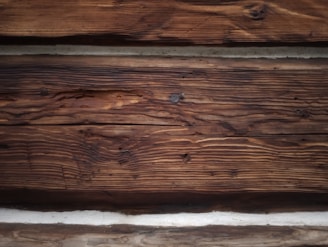Rough-textured wooden logs with various shades from dark brown to light tan. The logs are stacked horizontally and display natural grain patterns along with some knots and minor cracks.