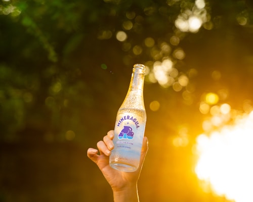 A hand is holding a clear glass bottle of sparkling water with a label visible against a backdrop of blurred, golden bokeh created by sunlight filtering through leaves.