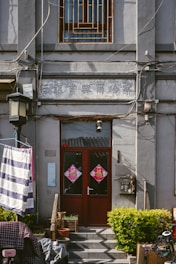 A building facade with two red doors decorated with traditional Chinese symbols. Above the doors, there is a weathered sign with Chinese writing. Various cables are attached to the building, and a street lamp is mounted on the wall. A striped cloth is hanging on the left side, and there are potted plants and a small bush near the entrance.