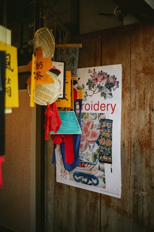 A wooden wall adorned with a colorful poster and various hanging decorations. The poster features floral embroidery designs, and traditional Chinese characters and imagery are present. Brightly colored fabrics in red, blue, and aqua green hang in front, adding vibrancy.