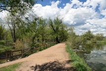 A serene pathway winds through lush greenery, bordered by a wooden fence. Tall trees with fresh green leaves arch over the path on one side, while a calm body of water reflects the cloudy blue sky on the other. The scene is peaceful and inviting, evoking a sense of calm and connection with nature.