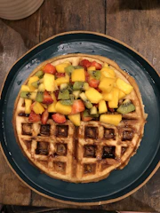A colorful waffle topped with fresh fruits, sauces, and sprinkles on a rustic wooden table.