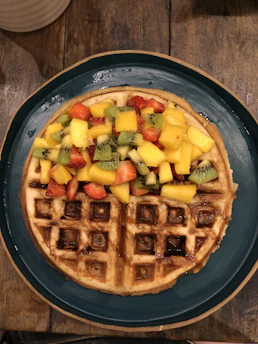 A colorful waffle topped with fresh fruits, sauces, and sprinkles on a rustic wooden table.