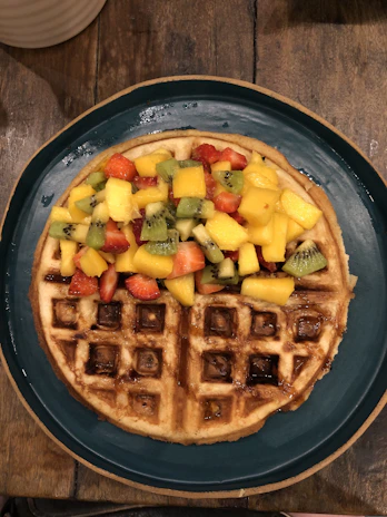 Close-up of a colorful, custom-designed waffle topped with fresh fruit and drizzled sauces on a rustic wooden table.
