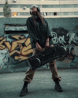 A person wearing sunglasses and headphones stands confidently in front of a graffiti-covered wall, holding a large boombox. The individual is dressed in casual, urban attire with a dark jacket, tan pants, and black boots.