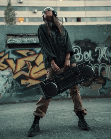 A person wearing sunglasses and headphones stands confidently in front of a graffiti-covered wall, holding a large boombox. The individual is dressed in casual, urban attire with a dark jacket, tan pants, and black boots.