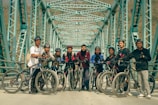 A group of people standing with bicycles on an iron bridge with a geometric pattern of beams overhead. They are wearing casual sportswear and most are equipped with helmets. The atmosphere appears friendly and adventurous.