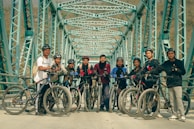A group of people standing with bicycles on an iron bridge with a geometric pattern of beams overhead. They are wearing casual sportswear and most are equipped with helmets. The atmosphere appears friendly and adventurous.