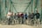 A group of people standing with bicycles on an iron bridge with a geometric pattern of beams overhead. They are wearing casual sportswear and most are equipped with helmets. The atmosphere appears friendly and adventurous.