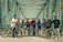 A group of people standing with bicycles on an iron bridge with a geometric pattern of beams overhead. They are wearing casual sportswear and most are equipped with helmets. The atmosphere appears friendly and adventurous.