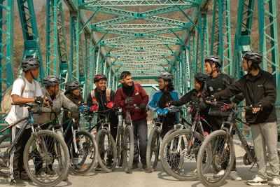 A group of nine cyclists with helmets and bikes are gathered on a teal-colored metal bridge. They appear to be engaged in conversation, with smiles and relaxed postures, suggesting a friendly interaction. The bridge structure creates a repetitive pattern in the background, enhancing the visual appeal of the scene.