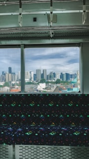 The image captures the interior of a train or subway car with a patterned seat in the foreground, looking out towards a city skyline through a large window. The skyline consists of numerous high-rise buildings under a partly cloudy sky.