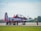 A small aircraft is stationed on an airstrip surrounded by a group of people wearing red uniforms. The plane has a red and white color scheme and a single propeller at the front. The airstrip is bordered by green grass, and a few trees are visible in the distance. A bird is flying in the blue sky above.