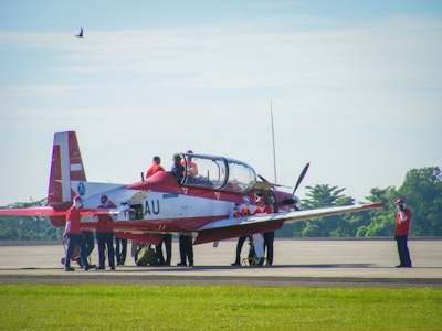 A small aircraft is stationed on an airstrip surrounded by a group of people wearing red uniforms. The plane has a red and white color scheme and a single propeller at the front. The airstrip is bordered by green grass, and a few trees are visible in the distance. A bird is flying in the blue sky above.