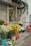 A storefront with various colorful flowers in pots arranged along the sidewalk. Sunflowers, lilies, and other flowers are displayed in blue, pink, and red containers. A glass door and window showcase additional plants and vases inside the shop. An awning provides shade, and a metal shutter is visible on the side.