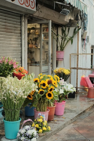 A storefront with various colorful flowers in pots arranged along the sidewalk. Sunflowers, lilies, and other flowers are displayed in blue, pink, and red containers. A glass door and window showcase additional plants and vases inside the shop. An awning provides shade, and a metal shutter is visible on the side.