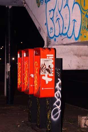 A row of four bright red public payphones attached to a wall covered with graffiti art in various colors. The phones have some yellow and white text and markings on them. The surrounding environment appears urban and slightly dimly lit, suggesting it might be nighttime.