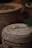 A close-up view of a terracotta pot with a rustic finish. On top of the pot, a small white flower and a dried leaf are carefully placed. The background features more earthen pots, slightly out of focus.