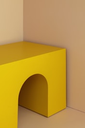 A minimalist composition featuring a geometric, rectangular object in bright yellow. The object has a curved cutout and is set against a muted beige background, creating a stark contrast between the vibrant yellow and the subtle backdrop.
