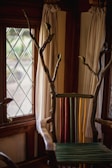 A uniquely designed chair with a back made of colorful vertical slats in green, yellow, and red. The arms and top are crafted from natural wood with branches extending upwards. The setting includes a window with diamond-patterned glass and light-colored curtains.