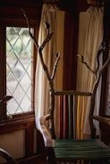 A uniquely designed chair with a back made of colorful vertical slats in green, yellow, and red. The arms and top are crafted from natural wood with branches extending upwards. The setting includes a window with diamond-patterned glass and light-colored curtains.