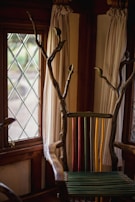 A uniquely designed chair with a back made of colorful vertical slats in green, yellow, and red. The arms and top are crafted from natural wood with branches extending upwards. The setting includes a window with diamond-patterned glass and light-colored curtains.