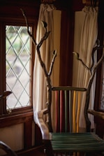 A uniquely designed chair with a back made of colorful vertical slats in green, yellow, and red. The arms and top are crafted from natural wood with branches extending upwards. The setting includes a window with diamond-patterned glass and light-colored curtains.