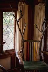 A uniquely designed chair with a back made of colorful vertical slats in green, yellow, and red. The arms and top are crafted from natural wood with branches extending upwards. The setting includes a window with diamond-patterned glass and light-colored curtains.