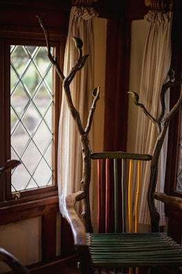 A uniquely designed chair with a back made of colorful vertical slats in green, yellow, and red. The arms and top are crafted from natural wood with branches extending upwards. The setting includes a window with diamond-patterned glass and light-colored curtains.