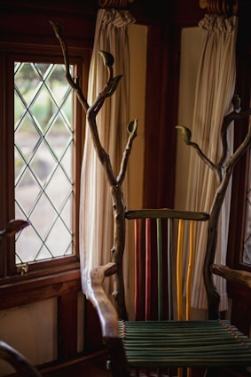 A uniquely designed chair with a back made of colorful vertical slats in green, yellow, and red. The arms and top are crafted from natural wood with branches extending upwards. The setting includes a window with diamond-patterned glass and light-colored curtains.