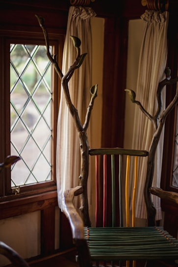A uniquely designed chair with a back made of colorful vertical slats in green, yellow, and red. The arms and top are crafted from natural wood with branches extending upwards. The setting includes a window with diamond-patterned glass and light-colored curtains.