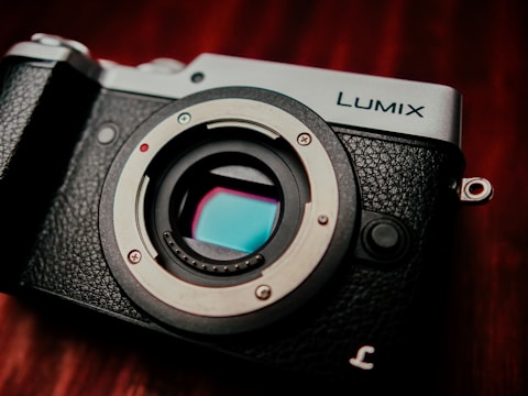 A close-up view of a black and silver camera with a visible sensor and the brand name 'Lumix' on the top. The textured surface of the camera body contrasts with the smooth, metallic lens mount.