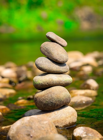 A balanced stack of smooth, rounded stones on a riverbed with a blurred background of green foliage. The stones are arranged vertically, creating a sense of harmony and equilibrium.