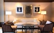 A well-furnished living room features a beige sofa with two light pillows, flanked by two elegant floor lamps on dark side tables. In front of the sofa, there is a round coffee table with a decorative geometric pattern, surrounded by two armchairs with striped upholstery. The walls are adorned with two framed artworks featuring intricate designs and a central abstract piece.