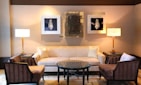 A well-furnished living room features a beige sofa with two light pillows, flanked by two elegant floor lamps on dark side tables. In front of the sofa, there is a round coffee table with a decorative geometric pattern, surrounded by two armchairs with striped upholstery. The walls are adorned with two framed artworks featuring intricate designs and a central abstract piece.