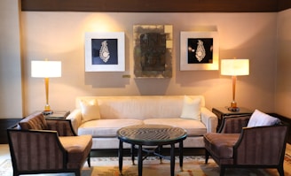 A well-furnished living room features a beige sofa with two light pillows, flanked by two elegant floor lamps on dark side tables. In front of the sofa, there is a round coffee table with a decorative geometric pattern, surrounded by two armchairs with striped upholstery. The walls are adorned with two framed artworks featuring intricate designs and a central abstract piece.