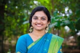 A woman is smiling warmly while wearing a traditional blue and green sari adorned with intricate patterns. Her earrings are large and decorative, and she has flowers in her hair. The background is lush and green, suggesting a natural or garden setting.