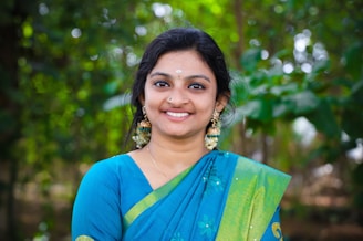 A woman is smiling warmly while wearing a traditional blue and green sari adorned with intricate patterns. Her earrings are large and decorative, and she has flowers in her hair. The background is lush and green, suggesting a natural or garden setting.