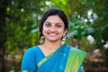 A woman is smiling warmly while wearing a traditional blue and green sari adorned with intricate patterns. Her earrings are large and decorative, and she has flowers in her hair. The background is lush and green, suggesting a natural or garden setting.