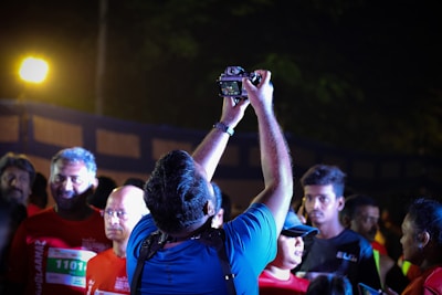 A group of people is gathered at an event, with one individual holding a camera above his head as if capturing a selfie. Several people around are wearing running attire with bib numbers, suggesting a marathon or sports event setting. The lighting is bright and focused, highlighting faces against a darker background.