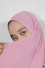 A close-up of a person's face partially covered by a pink headscarf, with expressive eyes prominently visible. The fabric drapes gracefully, creating a soft, elegant look.