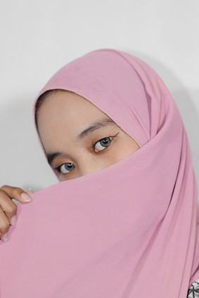 A close-up of a person's face partially covered by a pink headscarf, with expressive eyes prominently visible. The fabric drapes gracefully, creating a soft, elegant look.