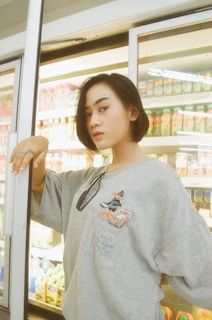 A person with short hair stands in front of refrigerators in a store, wearing a gray sweatshirt with a cartoon character print and sunglasses hanging from the neckline. The background shows various beverages in colorful bottles and cartons.