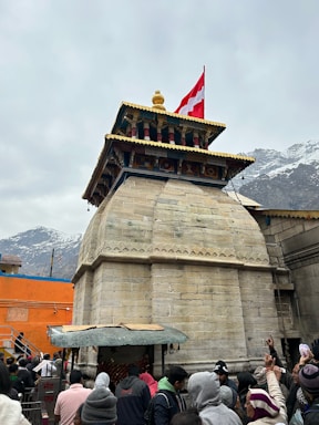 A stone temple structure with intricate architectural details stands prominently. A red and white flag waves majestically at the top. Surrounding the temple are snow-covered mountains that add a serene backdrop. The scene includes a crowd of people in various clothing, indicating a gathering or event.