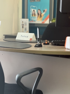 A desk setup with a computer monitor, a pen in a stand, a keyboard, and a mouse on a mat. There is a sign with the word 'Tutup' on the desk. In the background, there is a blurred poster with a smiling person.
