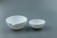 Two white ceramic bowls with pouring spouts sit side by side against a muted gray background. The bowls differ in size, with the larger one on the left and the smaller one on the right.