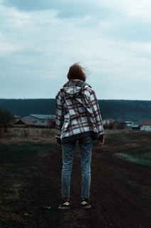 A person stands facing away in an outdoor setting, wearing a plaid jacket and jeans. The environment appears rural, with a few houses visible in the background and overcast skies above. The landscape is composed of grassy fields and a dirt path.