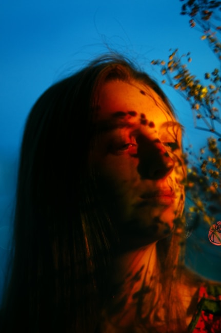 A close-up portrait of a female with long hair, bathed in warm, golden light. Shadows of foliage are cast on her face, with a blurred blue background adding contrast and depth. The image conveys a sense of tranquility and introspection.