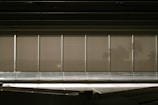 A row of vertical window blinds partially pulled down, with sunlight casting shadows and creating a warm, subdued atmosphere. In the background, faint silhouettes of palm trees can be seen through the blinds.