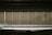 A row of vertical window blinds partially pulled down, with sunlight casting shadows and creating a warm, subdued atmosphere. In the background, faint silhouettes of palm trees can be seen through the blinds.